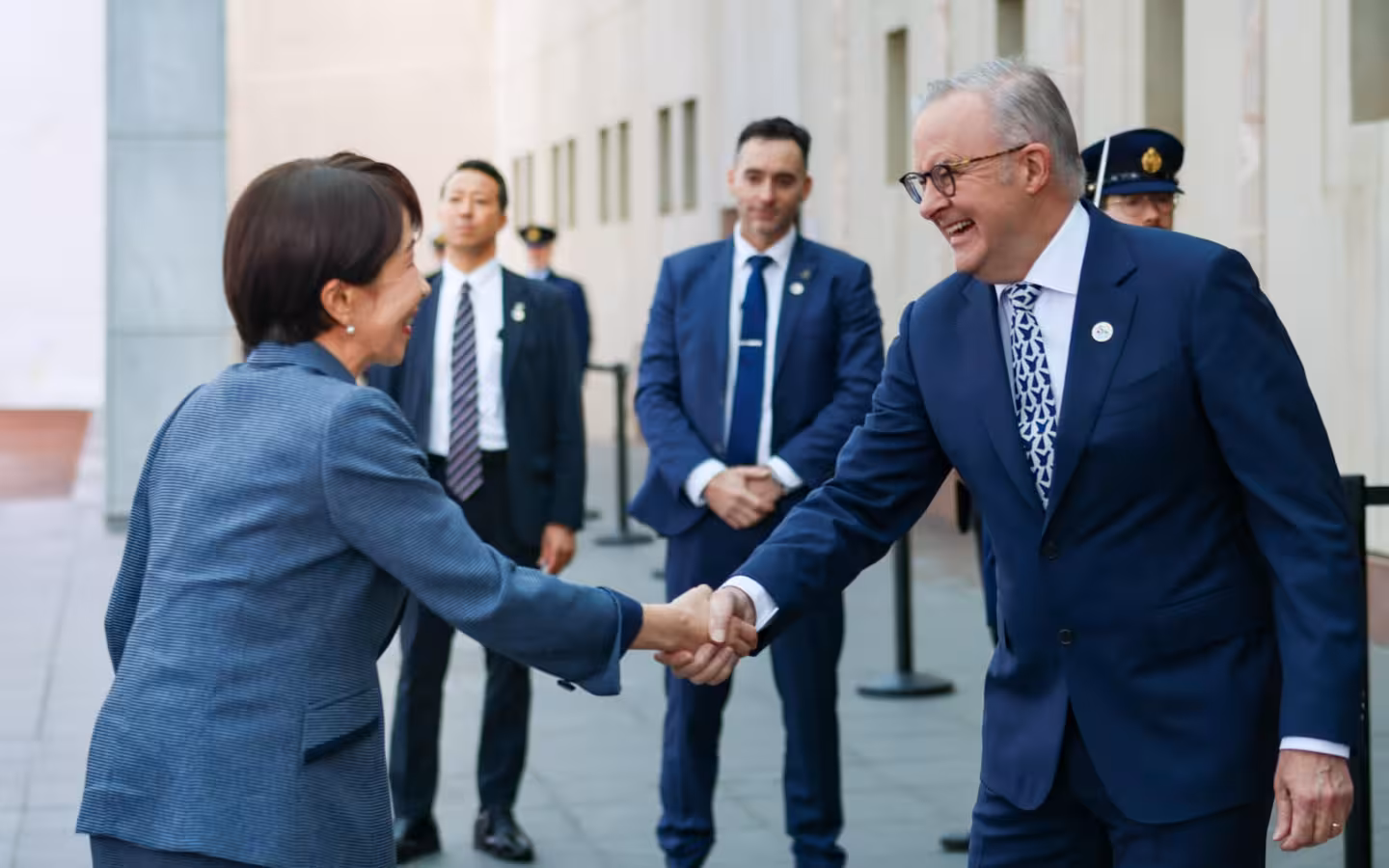 Two political figures shaking hands outdoors with several security guards standing behind them