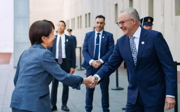 Two political figures shaking hands outdoors with several security guards standing behind them