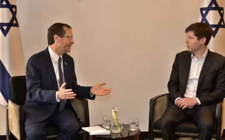 Isaac Herzog and Sam Altman sitting in armchairs talking, with Israeli flags behind them