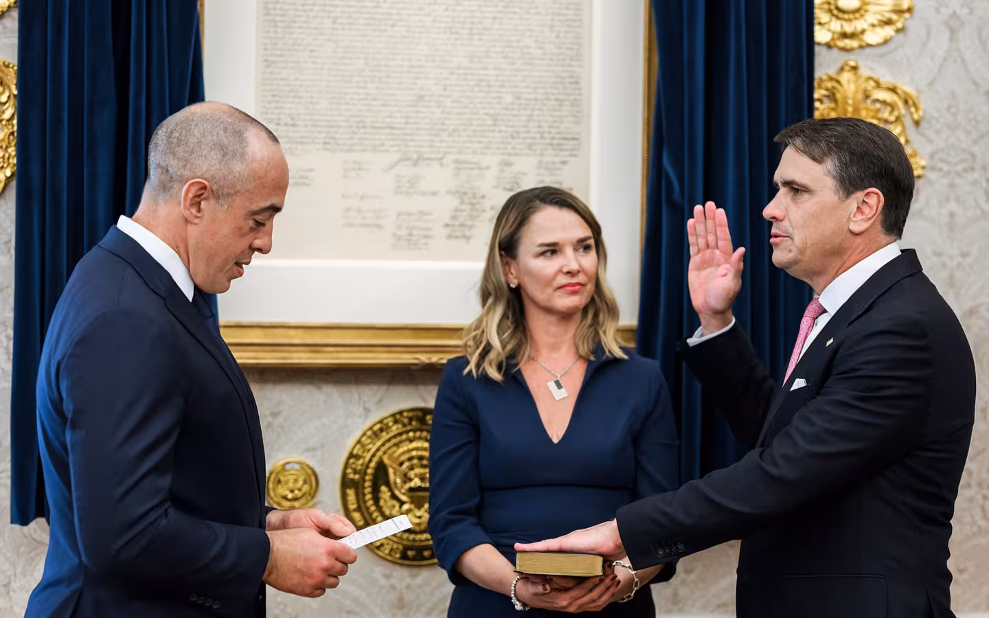 Three people indoors during a swearing in ceremony with a framed document behind them.
