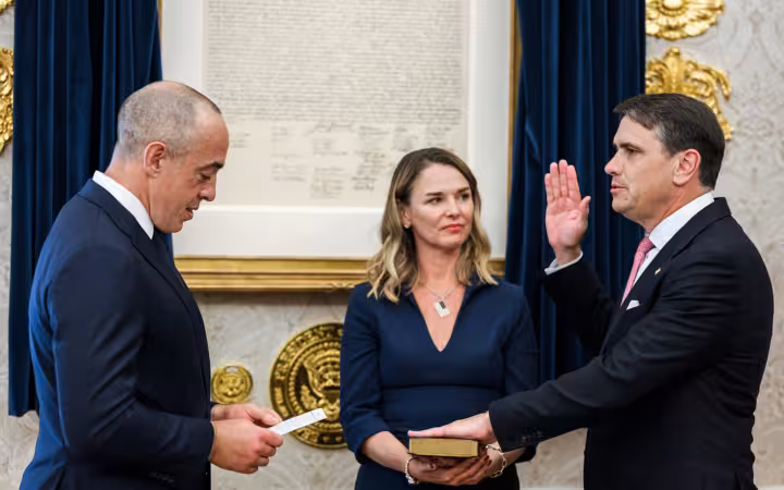 Three people indoors during a swearing in ceremony with a framed document behind them.