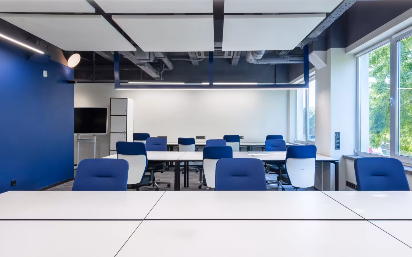 Modern office workspace with desks, chairs, large window, and monitor