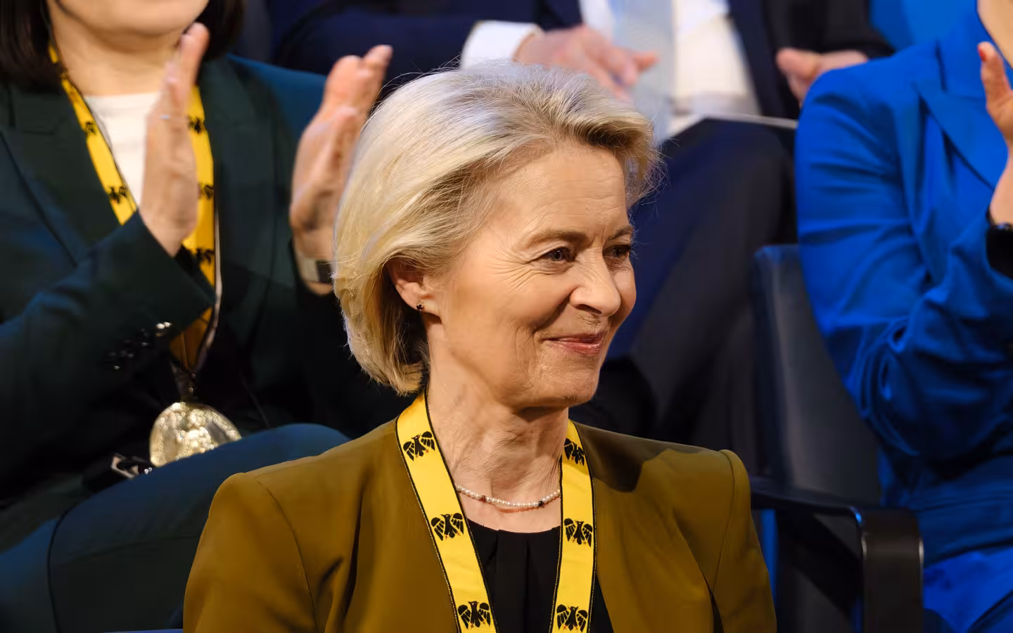 Ursula von der Leyen sitting in an audience during an event, smiling.