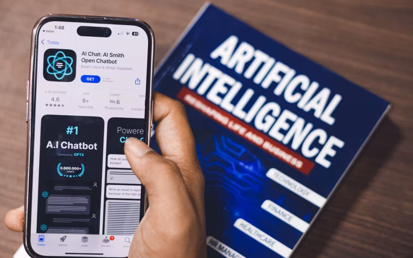 A hand holds a smartphone displaying an AI app beside an artificial intelligence book.