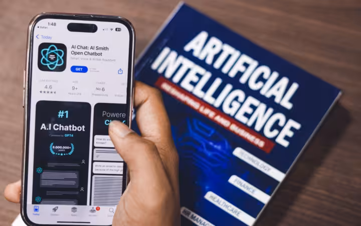A hand holds a smartphone displaying an AI app beside an artificial intelligence book.