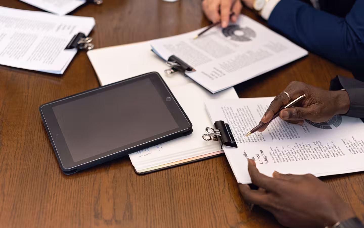 Hands review documents beside a dark tablet on a meeting table.