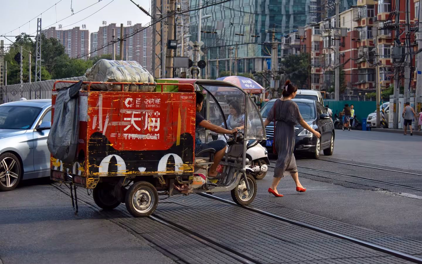 A Tmall-branded parcel trike crosses a busy Beijing street beside a pedestrian.