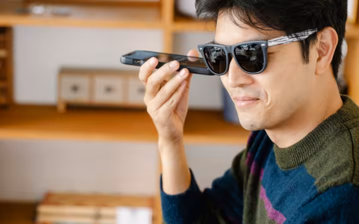 A man in sunglasses holds a smartphone near his mouth indoors.