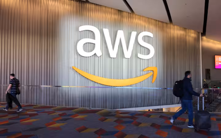 A large AWS sign on a conference wall as attendees walk past