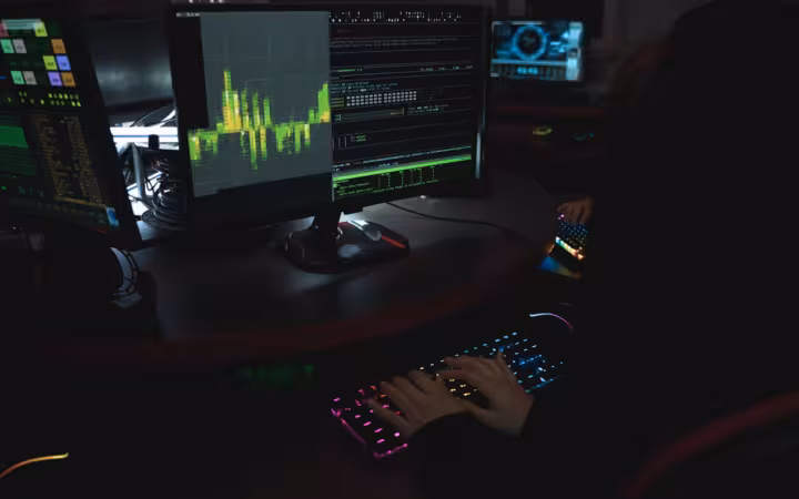 Hands type on a glowing keyboard beside multiple data-filled monitors.