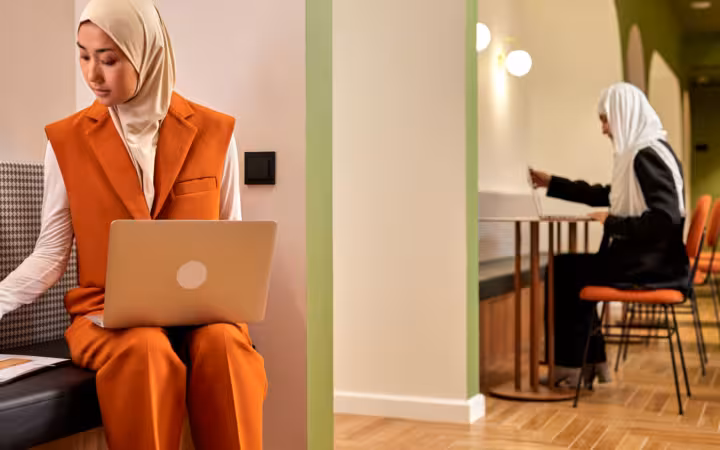 Two businesswomen in hijabs work on laptops in a modern office.