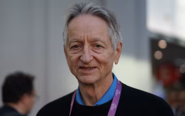 Geoffrey Hinton faces the camera with blurred conference attendees behind him.