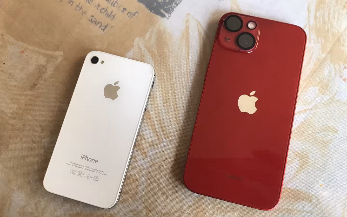 A red iPhone 13 and white iPhone 4S lie on patterned fabric.