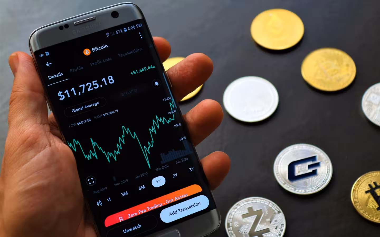 A hand holds a smartphone showing Bitcoin trading data beside cryptocurrency coins.