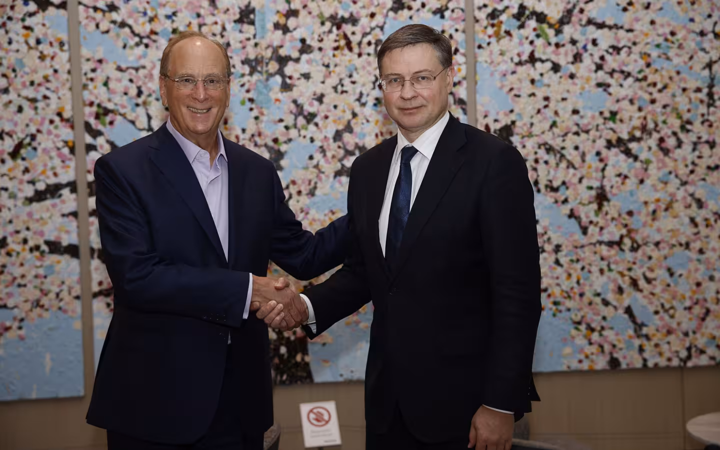 Larry Fink and Valdis Dombrovskis shake hands before a patterned wall.
