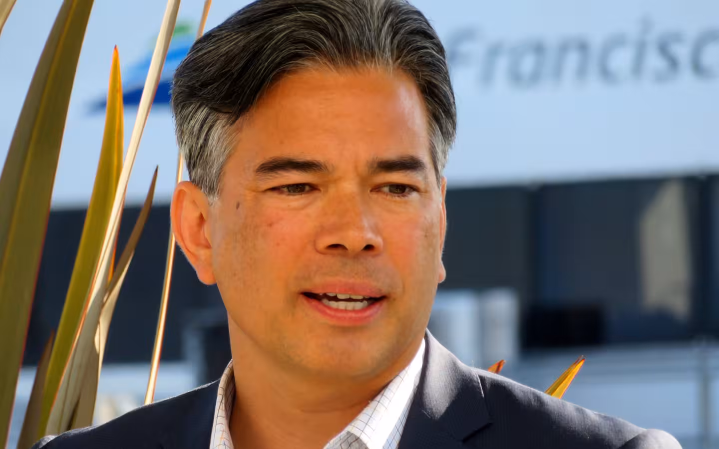 Rob Bonta speaking into a microphone outdoors, with plants and signage behind him