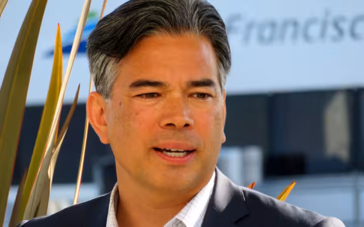 Rob Bonta speaking into a microphone outdoors, with plants and signage behind him