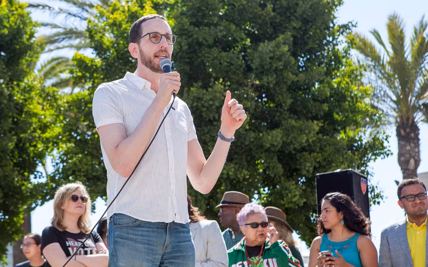 Scott Wiener speaks into a microphone outdoors as rally attendees stand behind him.
