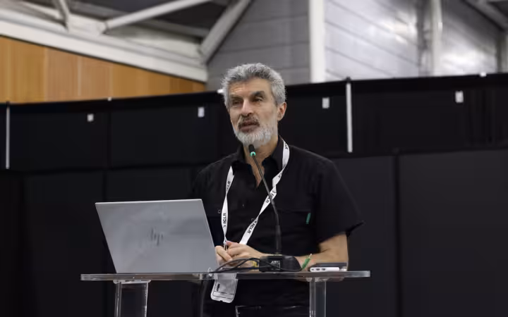Yoshua Bengio speaking into a microphone behind a laptop at ICLR 2025