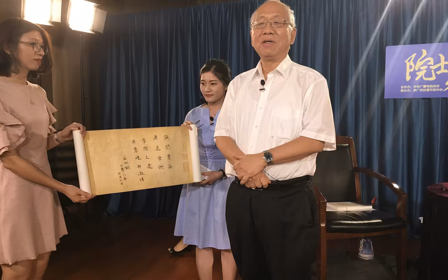 Shing-Tung Yau stands beside two women holding a calligraphy scroll in a studio.
