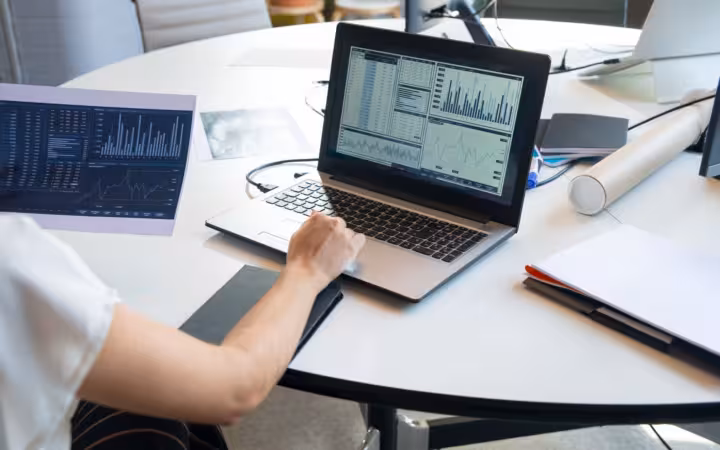 An office worker reviews financial charts on a laptop at a desk.