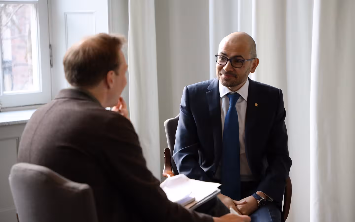Demis Hassabis seated opposite another man holding papers in a bright room