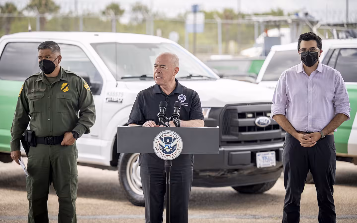 Alejandro Mayorkas speaks at a DHS lectern beside two officials and vehicles.