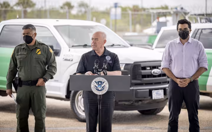 Alejandro Mayorkas speaks at a DHS lectern beside two officials and vehicles.