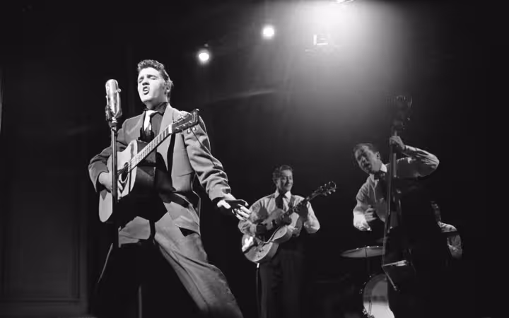 Elvis Presley sings into a microphone while playing guitar with bandmates.