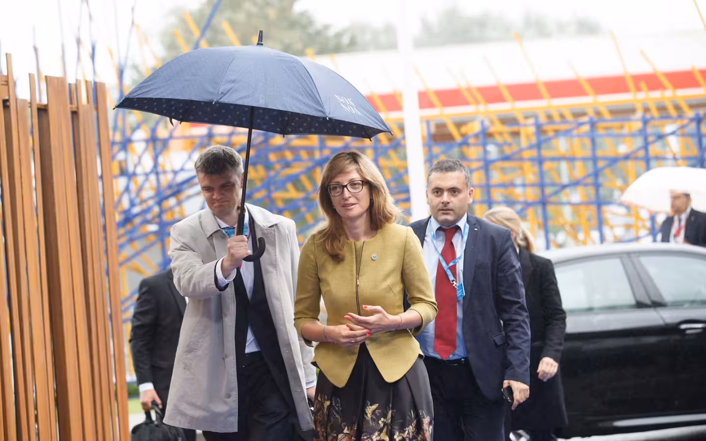 Ekaterina Zaharieva walks outdoors beneath an umbrella, accompanied by several people.