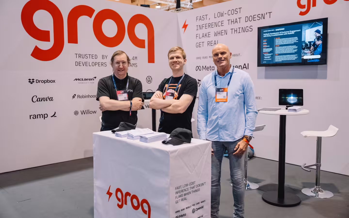 Three people pose behind a counter at a Groq-branded trade-show stand.