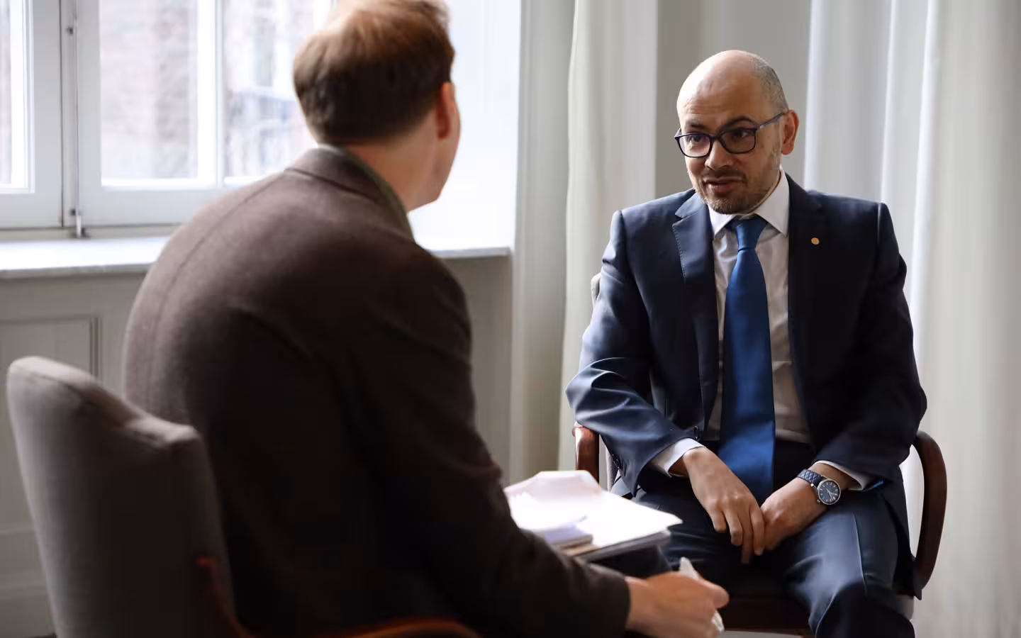 Demis Hassabis seated in conversation with another man seen from behind.