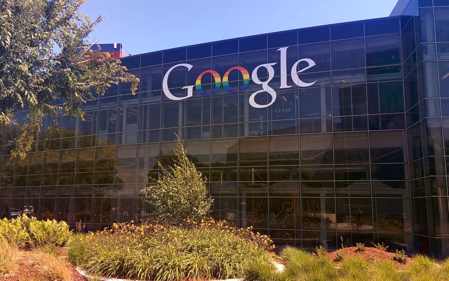 Googleplex glass facade displays a rainbow Google logo above landscaped grounds