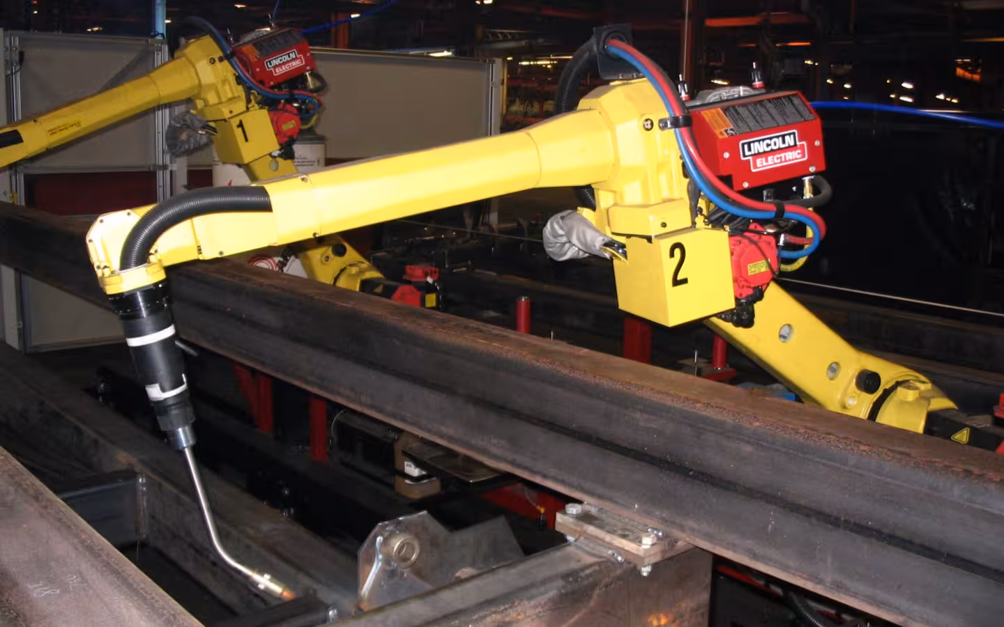 Two yellow Fanuc welding robots reach over steel beams inside a factory.