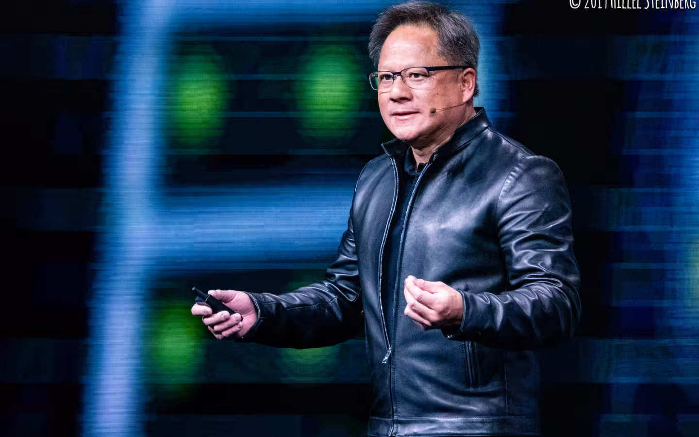 Jensen Huang speaks onstage in a black jacket against blue lighting.