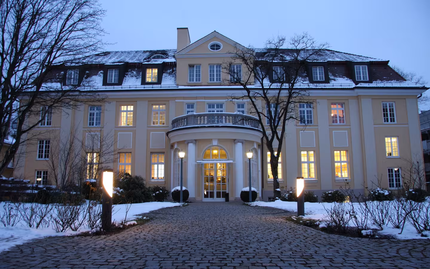 The ifo Institute’s snow-covered main building glows at dusk behind bare trees.