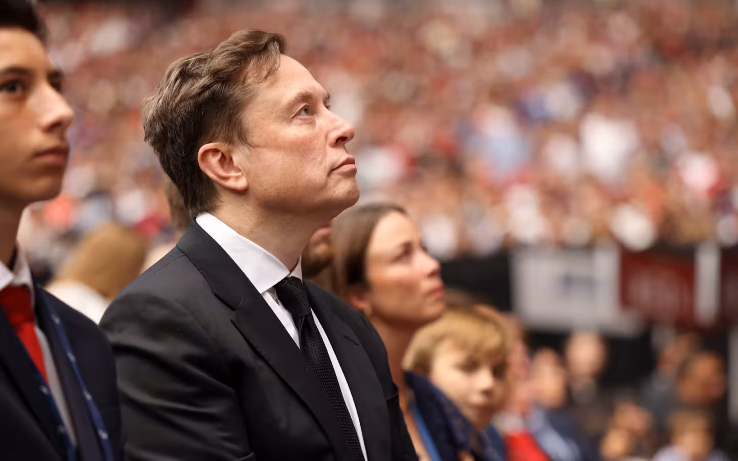 Elon Musk in a dark suit looks upward amid a crowded stadium.