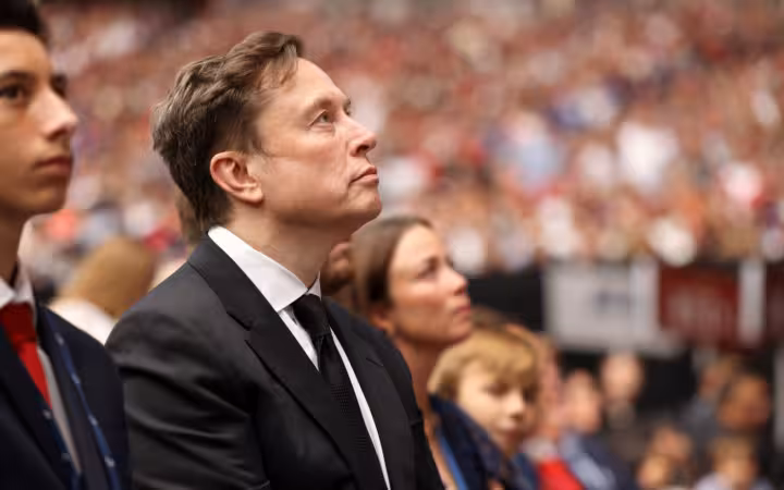 Elon Musk in a dark suit looks upward amid a crowded stadium.