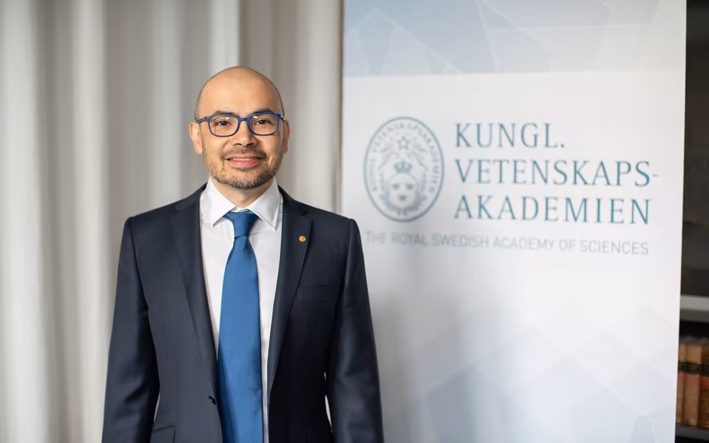 Demis Hassabis stands beside a Royal Swedish Academy of Sciences banner.