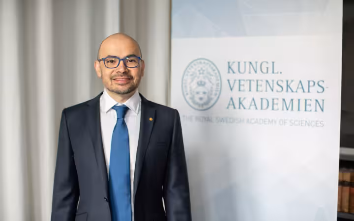 Demis Hassabis stands beside a Royal Swedish Academy of Sciences banner.