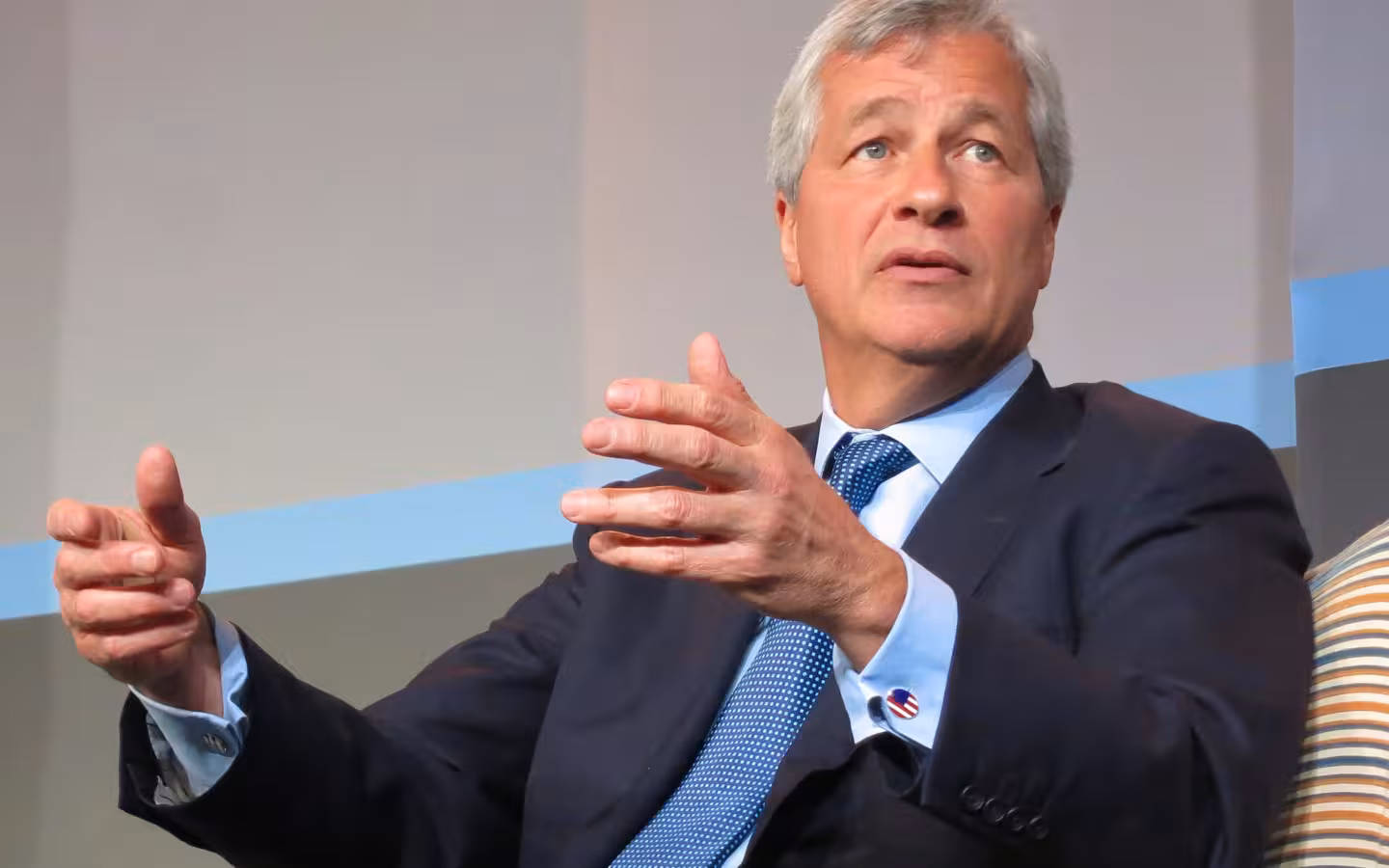 Jamie Dimon gestures while seated against a plain conference backdrop.
