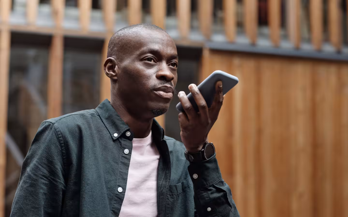 A man speaks toward a smartphone outdoors against a wooden building.