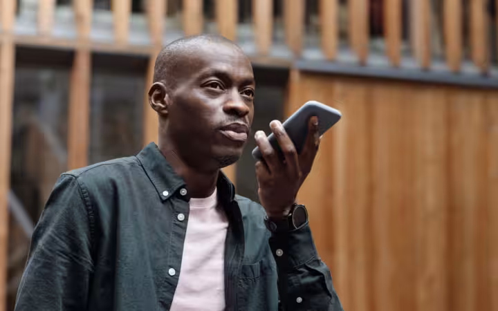 A man speaks toward a smartphone outdoors against a wooden building.