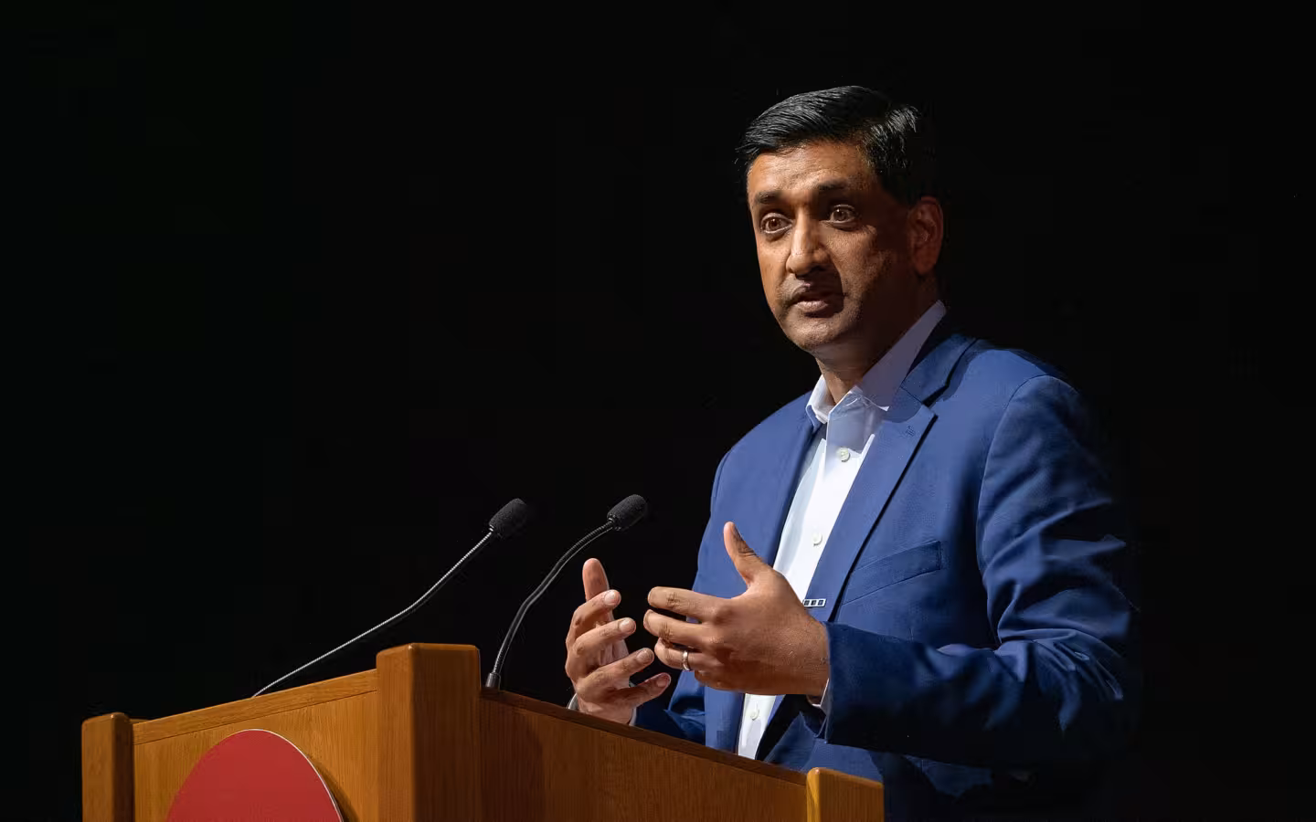 Congressman Ro Khanna speaking at a wooden podium with microphones in front.
