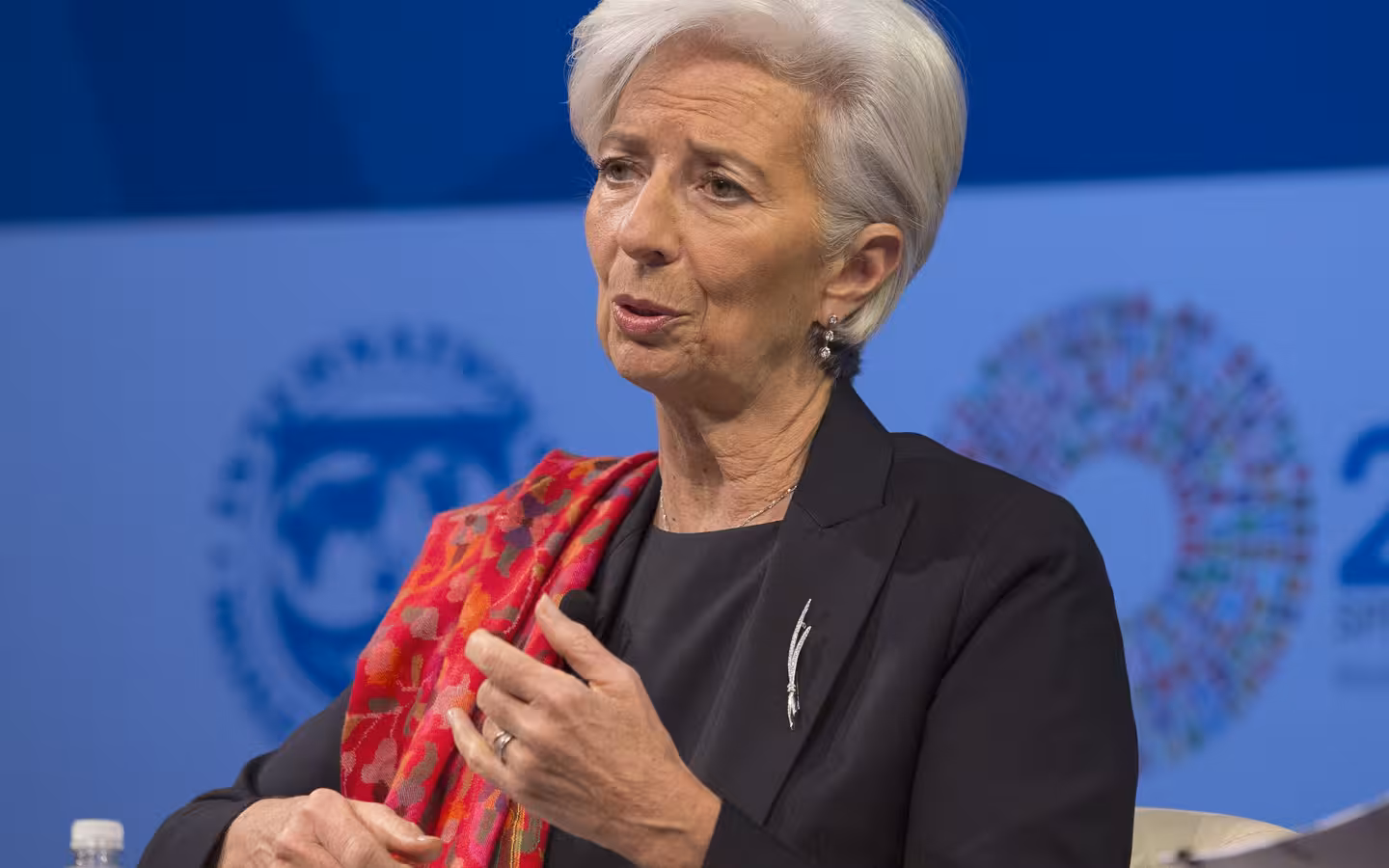 Christine Lagarde speaking and gesturing against a blue conference backdrop.