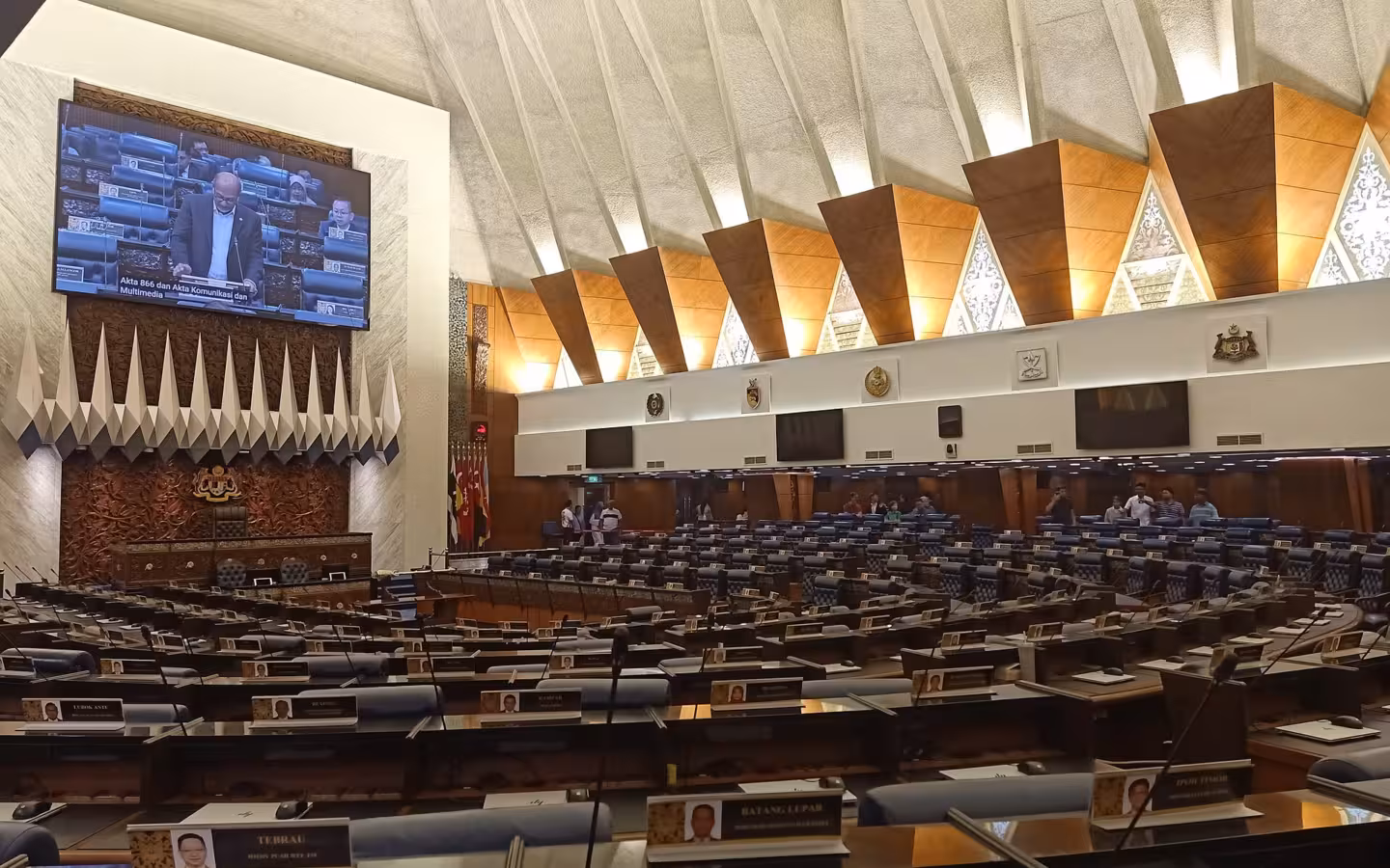 Dewan Rakyat chamber with rows of seats and a speaker on screen