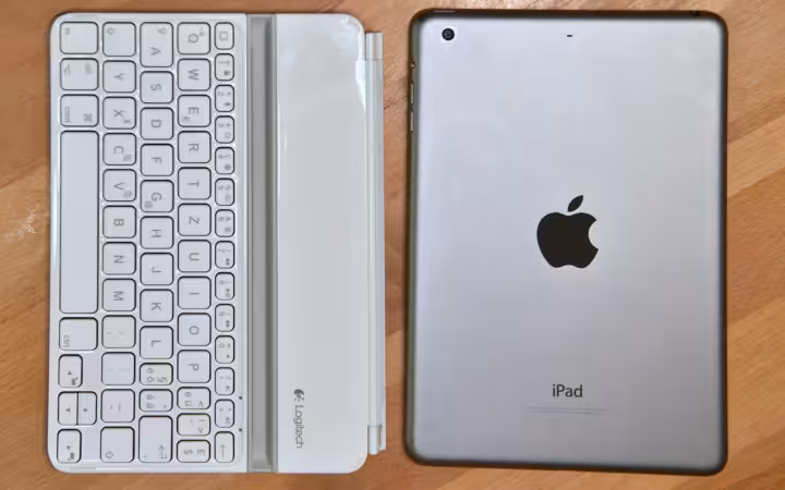 An Apple iPad mini beside a white Logitech keyboard on a wooden surface