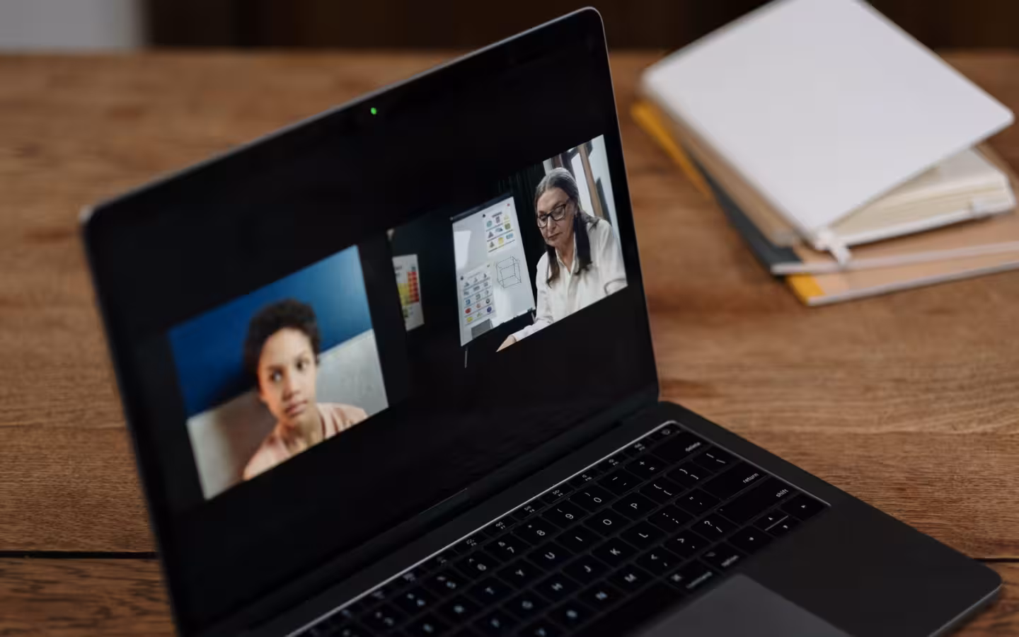 Two people appear on a laptop video call beside stacked notebooks.