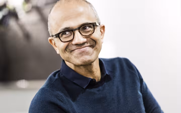 Microsoft CEO Satya Nadella smiling in glasses and a dark shirt.