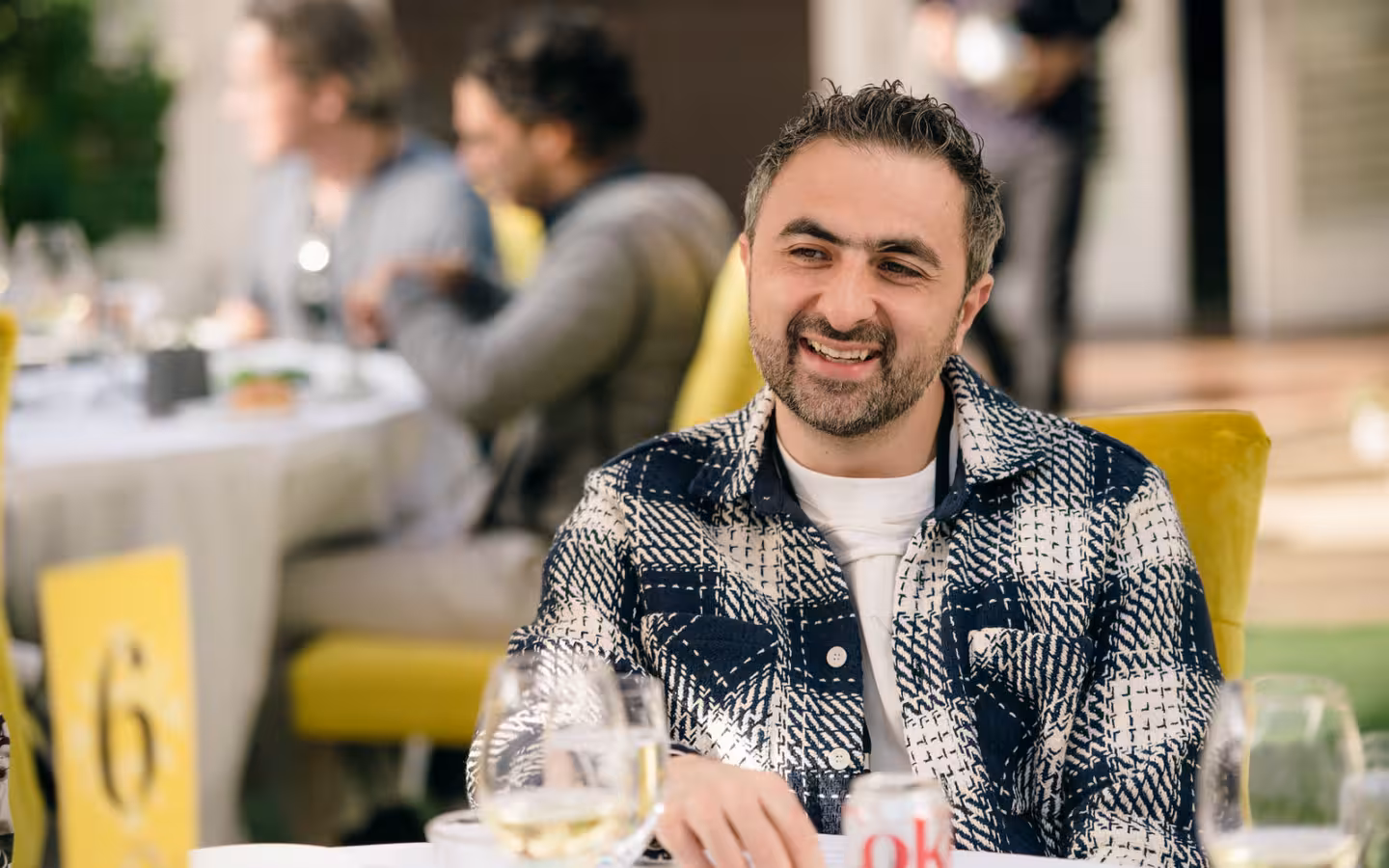 Mustafa Suleyman smiles at a dining table with people blurred behind him.